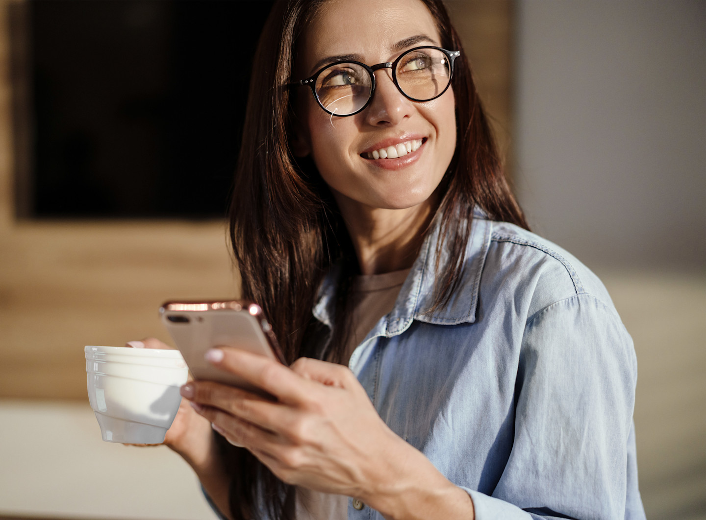 Frau mit Brille blickt lächelnd nach oben, in den Händen Smartphone und Kaffeetasse.